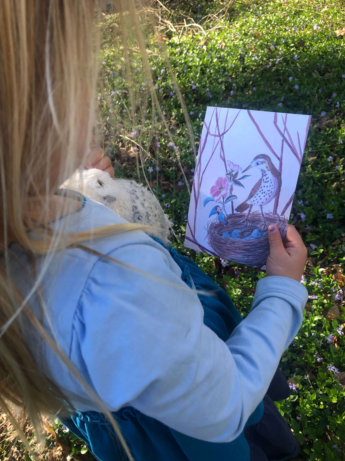 Young 4 year old holding illustration of a blue gnome holding a flower up to a singing thrush bird beside her on a nest. The child is also holding a toy owl. 