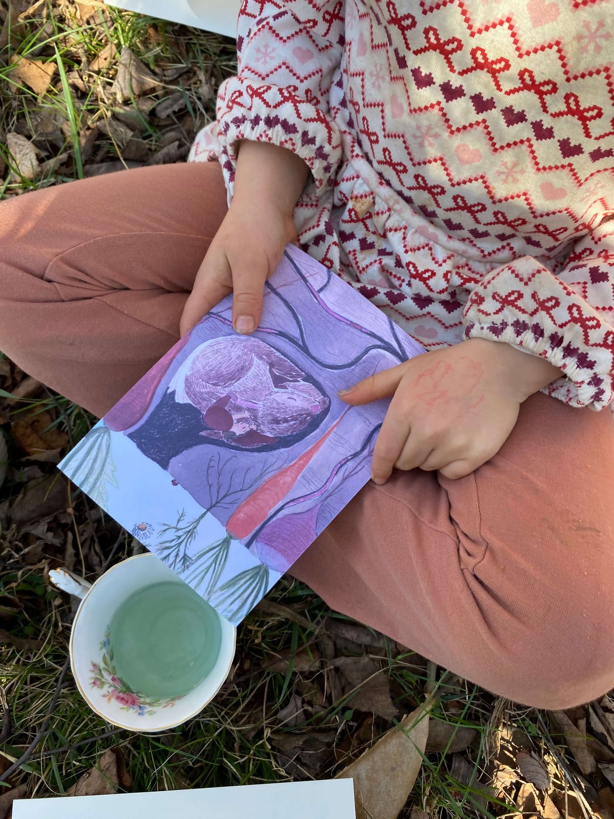 Six year old sitting on grass, crossed legs, holding a illustration of a red gnome asleep on a bunny underground surrounded by root vegetables. The child has a cup of blue-green tea beside her. 